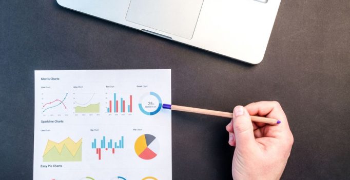 Hand holding pencil reviewing colorful data charts on desk with laptop.